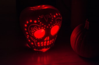 A carved pumpkin decorated with intricate patterns resembling a skull is illuminated from within by a red light, creating a glowing effect. The pumpkin is set against a dark background, enhancing the dramatic lighting and shadow play.