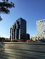 The image depicts a modern cityscape with a tall, contemporary office building featuring a glass facade and geometric design. In the foreground, there is a construction site with piles of earth and a metal barricade. Another building with a grid-like pattern of windows is visible on the right.
