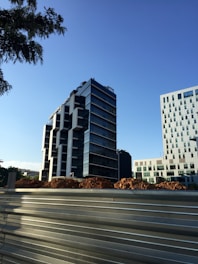 The image depicts a modern cityscape with a tall, contemporary office building featuring a glass facade and geometric design. In the foreground, there is a construction site with piles of earth and a metal barricade. Another building with a grid-like pattern of windows is visible on the right.