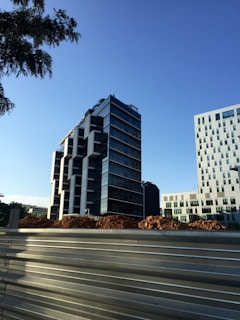 The image depicts a modern cityscape with a tall, contemporary office building featuring a glass facade and geometric design. In the foreground, there is a construction site with piles of earth and a metal barricade. Another building with a grid-like pattern of windows is visible on the right.