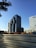 The image depicts a modern cityscape with a tall, contemporary office building featuring a glass facade and geometric design. In the foreground, there is a construction site with piles of earth and a metal barricade. Another building with a grid-like pattern of windows is visible on the right.