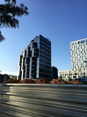 The image depicts a modern cityscape with a tall, contemporary office building featuring a glass facade and geometric design. In the foreground, there is a construction site with piles of earth and a metal barricade. Another building with a grid-like pattern of windows is visible on the right.
