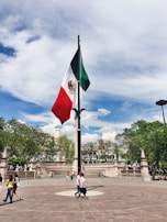 A large Mexican flag is prominently displayed in a public square surrounded by landscaped greenery and stone structures. People are casually strolling around the plaza, enjoying a sunny day. The sky is expansive with scattered clouds.