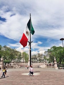 A large Mexican flag is prominently displayed in a public square surrounded by landscaped greenery and stone structures. People are casually strolling around the plaza, enjoying a sunny day. The sky is expansive with scattered clouds.