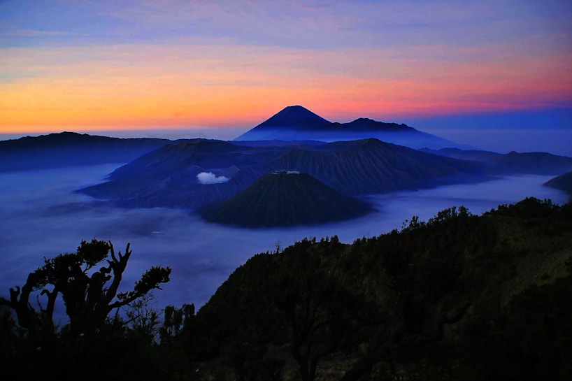 A breathtaking mountain landscape during sunrise, featuring layers of volcanic peaks shrouded in mist. The sky transitions from rich blue to vibrant orange and pink hues, creating a serene and majestic atmosphere. Dark silhouettes of trees are visible in the foreground, adding depth to the scene.