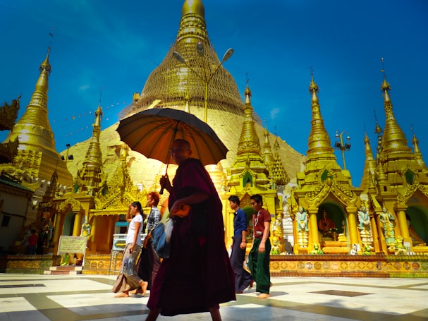 A group of people, including a monk holding an umbrella, walk barefoot around a golden pagoda under a clear blue sky. The pagoda is elaborate and richly decorated, with multiple spires and ornate carvings that shimmer in the sunlight.
