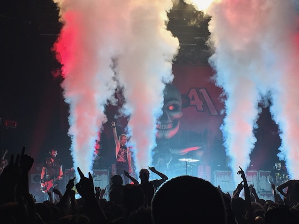 A dynamic concert scene with a band performing on stage surrounded by thick columns of smoke. The background features a large, ominous skull design with red accents. The crowd is visibly engaged, raising their arms in excitement as colored lights create an intense atmosphere.