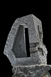 A textured stone sculpture with geometric cutouts, showcasing intricate craftsmanship and angular design against a black background. The sculpture is rugged yet meticulously shaped, with a blend of rough and smooth surfaces.