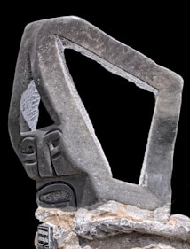 A modern, abstract stone sculpture with a geometric shape and a stylized face. The stone is rough-textured, with a prominent open, rectangular section in the upper part. The sculpture is mounted on a rocky base, and a small plaque is visible at the bottom.