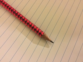 A sharpened pencil with a vibrant red and black checkered pattern rests on a page of a lined notebook. The pencil's wood is exposed at the tip, showing its graphite point ready for writing.