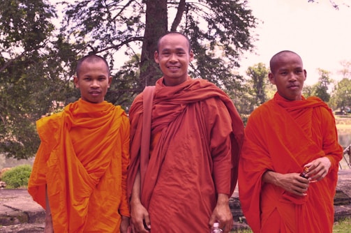 Three monks in vibrant orange robes stand together outdoors, with a background of trees and greenery. They are smiling gently, creating a serene and peaceful atmosphere.