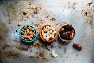 Three small bowls are arranged on a textured metal surface, each containing different types of nuts and dried fruits. The leftmost bowl, which is turquoise, holds small round tiger nuts. The middle bowl, orange in color, contains cashew nuts, and is positioned slightly lower than the other two. On the right, a brown bowl is filled with a few dates. Scattered around the bowls are additional nuts and dates that add to the rustic composition.