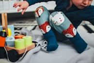 A young child dressed in patterned leggings with playful animal designs, including a fox and a rabbit, is sitting on a soft blanket. Nearby, there is a wooden toy with colorful cylindrical blocks in shades of blue, yellow, and orange.