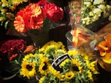 A vibrant assortment of flower bouquets in various colors including red, orange, yellow, and white. Prominently featured are sunflowers, marked with a small chalkboard sign indicating their price. The arrangement includes other flowers such as gerbera daisies and calla lilies in cellophane wrapping.