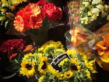 A vibrant assortment of flower bouquets in various colors including red, orange, yellow, and white. Prominently featured are sunflowers, marked with a small chalkboard sign indicating their price. The arrangement includes other flowers such as gerbera daisies and calla lilies in cellophane wrapping.