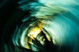 Vivid swirling patterns of light and color create a dynamic motion effect, blending shades of turquoise, yellow, and black. The abstract composition resembles a vortex or whirlpool.