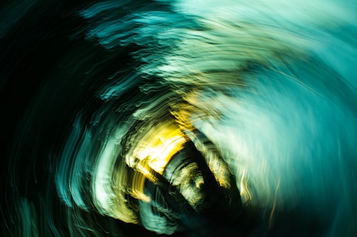 Vivid swirling patterns of light and color create a dynamic motion effect, blending shades of turquoise, yellow, and black. The abstract composition resembles a vortex or whirlpool.
