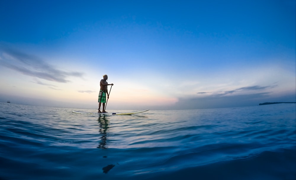 man standing on white paddle board holding paddle on body of water under blue sky