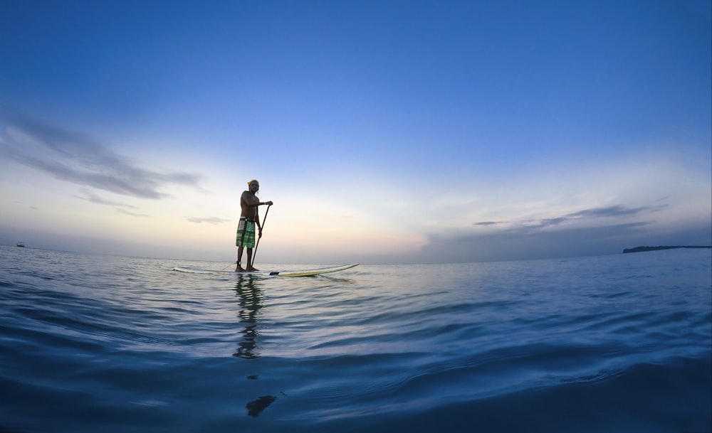 uomo in piedi sulla tavola da paddle bianca che tiene la pagaia sullo specchio d'acqua sotto il cielo blu