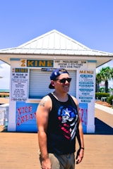 A person in a sleeveless shirt and cap is smiling, standing outside a surf and paddleboard rental kiosk. The kiosk features signs advertising ice cream and various water sports activities. Palm trees and a clear blue sky are visible in the background.