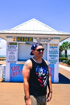 A person in a sleeveless shirt and cap is smiling, standing outside a surf and paddleboard rental kiosk. The kiosk features signs advertising ice cream and various water sports activities. Palm trees and a clear blue sky are visible in the background.