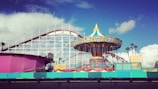 A vibrant amusement park scene featuring a colorful carousel with a decorative top. In the background, a classic white and red roller coaster extends into the sky. Blue sky with scattered white clouds creates a cheerful atmosphere. Brightly colored barriers and structures, including vibrant pink and turquoise hues, frame the attractions.