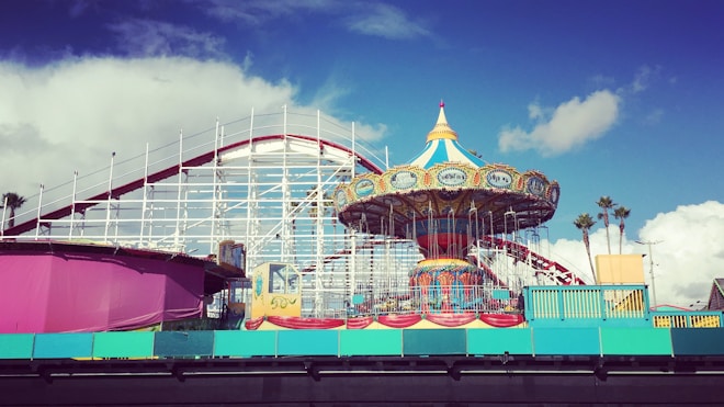 A vibrant amusement park scene featuring a colorful carousel with a decorative top. In the background, a classic white and red roller coaster extends into the sky. Blue sky with scattered white clouds creates a cheerful atmosphere. Brightly colored barriers and structures, including vibrant pink and turquoise hues, frame the attractions.