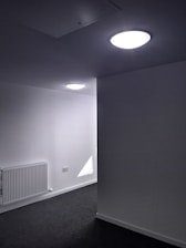 A minimalist interior space with gray carpeted flooring and bright white walls, featuring two ceiling lights. A radiator is installed on one of the walls, and an electrical outlet is visible.