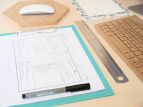 A neatly arranged workspace with a wooden keyboard, a metal ruler, and a mouse on a cork pad. A clipboard holds a document with a grid pattern, alongside a pen. The surface is light wood, and there is a decorative notepad visible in the background.