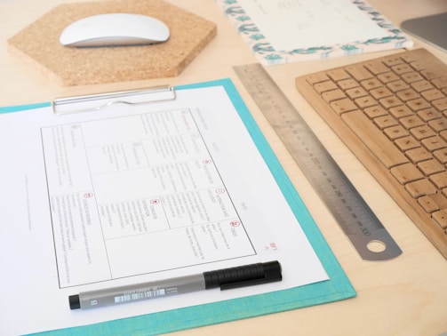 A neatly arranged workspace with a wooden keyboard, a metal ruler, and a mouse on a cork pad. A clipboard holds a document with a grid pattern, alongside a pen. The surface is light wood, and there is a decorative notepad visible in the background.