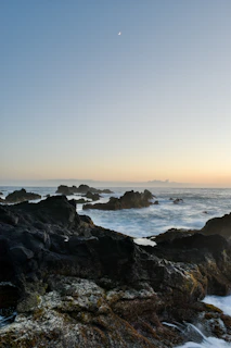 A wide shot of a rugged coastline at dusk, waves crashing against jagged rocks under a pastel sky.