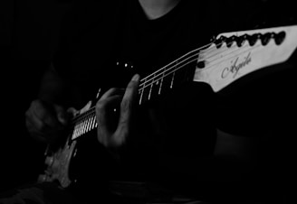 A person is playing an electric guitar. The image is captured in black and white, highlighting the musician's fingers on the fretboard and the guitar headstock with the brand logo visible. The focus is on the hand's positioning and the strings of the guitar.