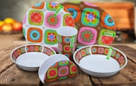 A set of brightly colored ceramic dishes with a floral and geometric pattern, including plates, bowls, a cup, and a napkin holder. They are placed on a wooden table, and the background is softly blurred with indistinct warm colors, suggesting a cozy kitchen setting.