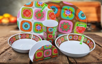 A set of brightly colored ceramic dishes with a floral and geometric pattern, including plates, bowls, a cup, and a napkin holder. They are placed on a wooden table, and the background is softly blurred with indistinct warm colors, suggesting a cozy kitchen setting.