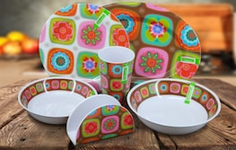 A set of brightly colored ceramic dishes with a floral and geometric pattern, including plates, bowls, a cup, and a napkin holder. They are placed on a wooden table, and the background is softly blurred with indistinct warm colors, suggesting a cozy kitchen setting.