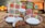 A set of brightly colored ceramic dishes with a floral and geometric pattern, including plates, bowls, a cup, and a napkin holder. They are placed on a wooden table, and the background is softly blurred with indistinct warm colors, suggesting a cozy kitchen setting.