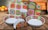 A set of brightly colored ceramic dishes with a floral and geometric pattern, including plates, bowls, a cup, and a napkin holder. They are placed on a wooden table, and the background is softly blurred with indistinct warm colors, suggesting a cozy kitchen setting.