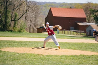 A baseball player in a red and white uniform is pitching on a field with a grassy outfield and a dirt pitcher's mound. In the background, there are trees and a red barn-like structure. Another player, wearing a blue helmet, is positioned in the outfield.