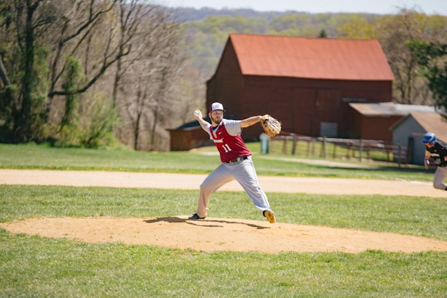 A baseball player in a red and white uniform is pitching on a field with a grassy outfield and a dirt pitcher's mound. In the background, there are trees and a red barn-like structure. Another player, wearing a blue helmet, is positioned in the outfield.