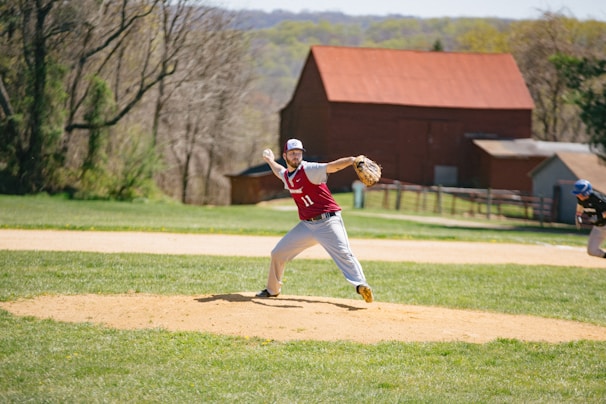 A baseball player in a red and white uniform is pitching on a field with a grassy outfield and a dirt pitcher's mound. In the background, there are trees and a red barn-like structure. Another player, wearing a blue helmet, is positioned in the outfield.