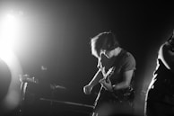 A black and white image of a musician playing an electric guitar. The person has medium-length hair and is wearing a short sleeve shirt. Intense lighting from the left side creates a dramatic contrast, casting shadows and adding to the dynamic atmosphere of the performance.