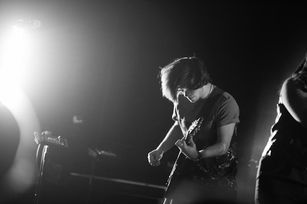 A black and white image of a musician playing an electric guitar. The person has medium-length hair and is wearing a short sleeve shirt. Intense lighting from the left side creates a dramatic contrast, casting shadows and adding to the dynamic atmosphere of the performance.