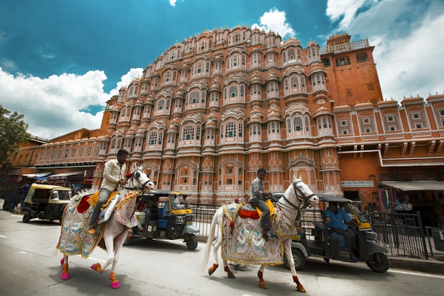 A grand, detailed facade of a pink sandstone palace stands prominently, showcasing an intricate array of windows and balconies. In the foreground, two men ride elaborately decorated horses along a street, with vibrant textiles adorning the animals. There are also auto-rickshaws and other street elements visible.
