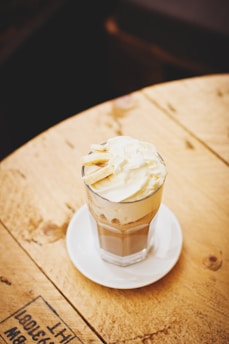 A glass of creamy iced coffee topped with whipped cream, served on a white saucer. The wooden table surface underneath adds a rustic feel to the setting.