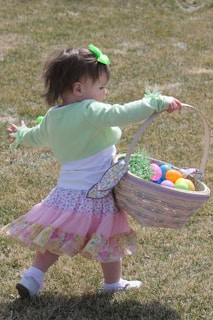 A young child with a green bow in their hair holds a wicker basket filled with colorful Easter eggs and grass. The child is dressed in a light green shirt and a pink, frilly skirt, and walks on a patch of grass.