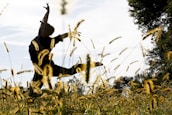 A silhouette of a person gracefully dancing in a field of tall grasses, against a backdrop of a clear sky. The sunlight creates a bright and serene atmosphere, illuminating the grass and casting playful shadows.