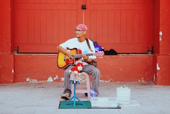An older man wearing a red bandana and glasses is seated while playing an acoustic guitar. He is in front of a large red door or wall. He is dressed in a white t-shirt and striped pants, sitting on a pink chair with various items around him, including a red toy, a plastic container, and a white bag.