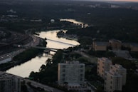 An aerial view of an urban landscape featuring a river surrounded by dense green foliage. Several bridges span the waterway, and a prominent neoclassical building is visible in the right mid-ground. Residential and commercial buildings align the lower section of the scene. Traffic is visible on the nearby roads.