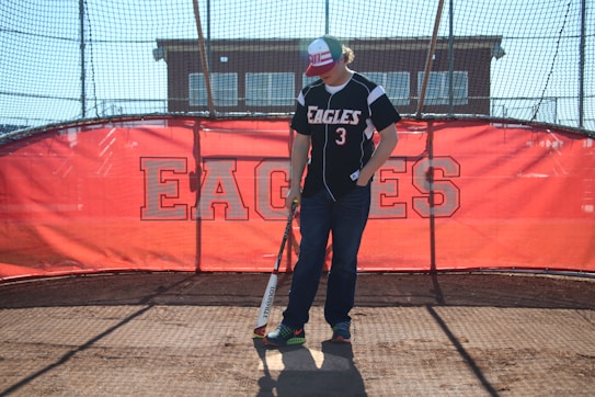A person wearing a black sports jersey with the word 'Eagles' stands in a batting cage. They hold a baseball bat and are surrounded by a red netting with 'EAGLES' printed on it. The background includes a building visible through the netting.