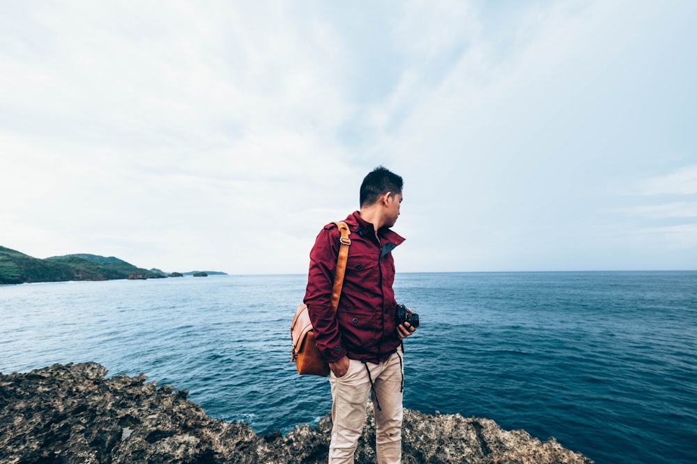 man wearing red zip-up jacket standing on top of rocky cliff beside body of water during daytime